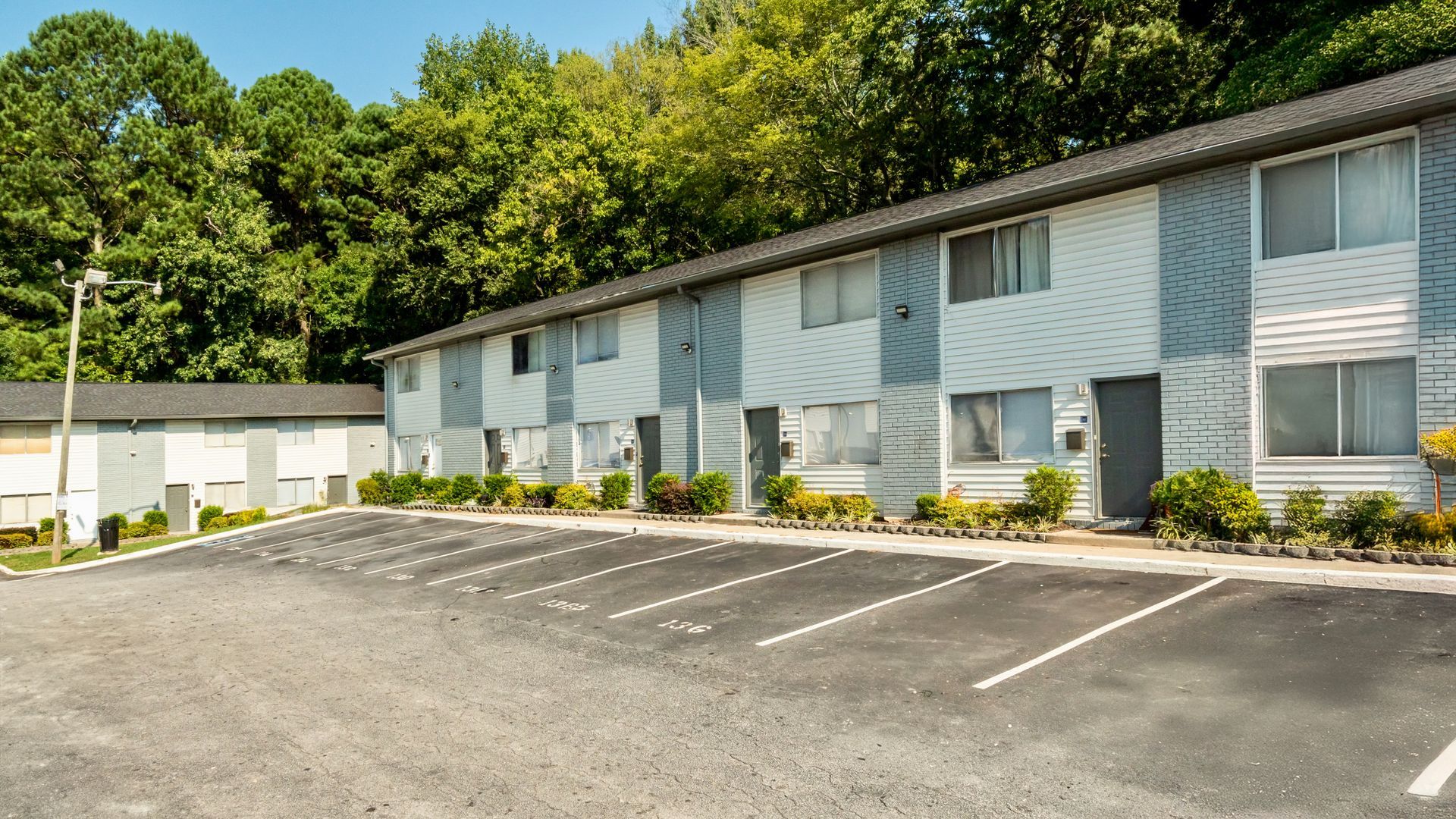 Apartment building with white siding and parking spaces in front, beside a tree-covered hillside
