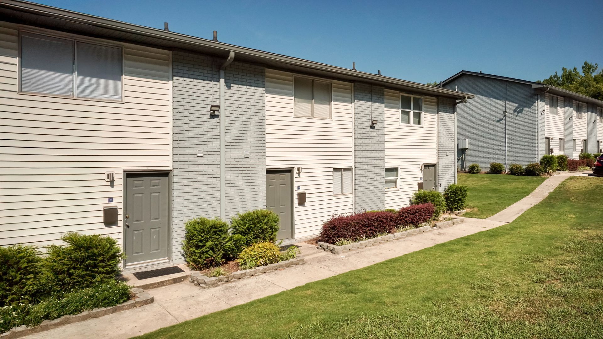 Row of beige apartment buildings with gray doors beside a sloping green lawn and sidewalk