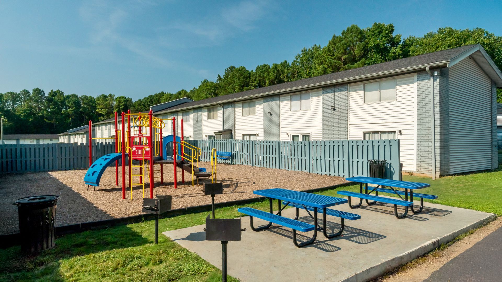 Playground with blue picnic tables beside a white building and a colorful play structure.