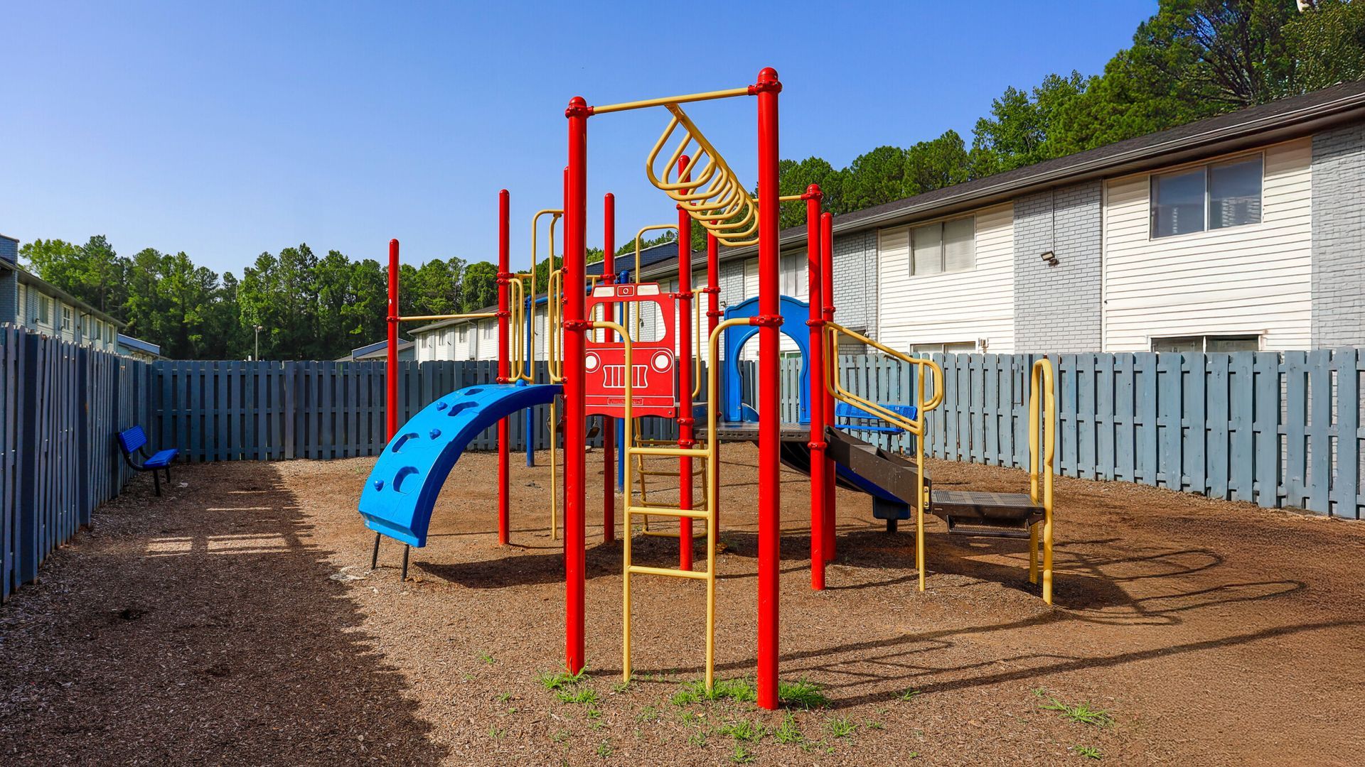 Empty playground with red climbing frame, blue slide, and yellow bars beside a building and fence.