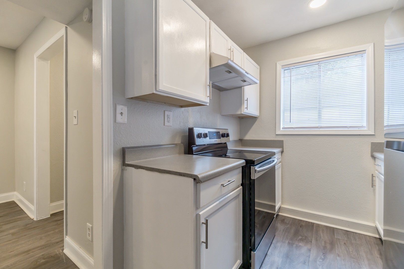 Small kitchen with white cabinets, stove, and window with blinds, in a bright apartment.
