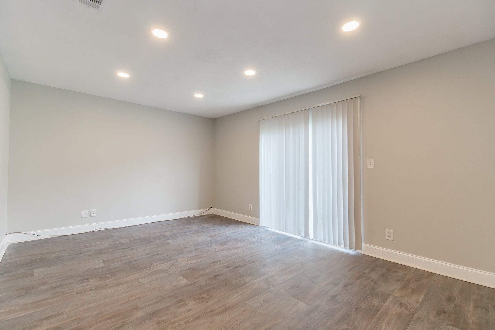 Empty beige room with wood flooring, recessed lights, and sliding glass door with vertical blinds