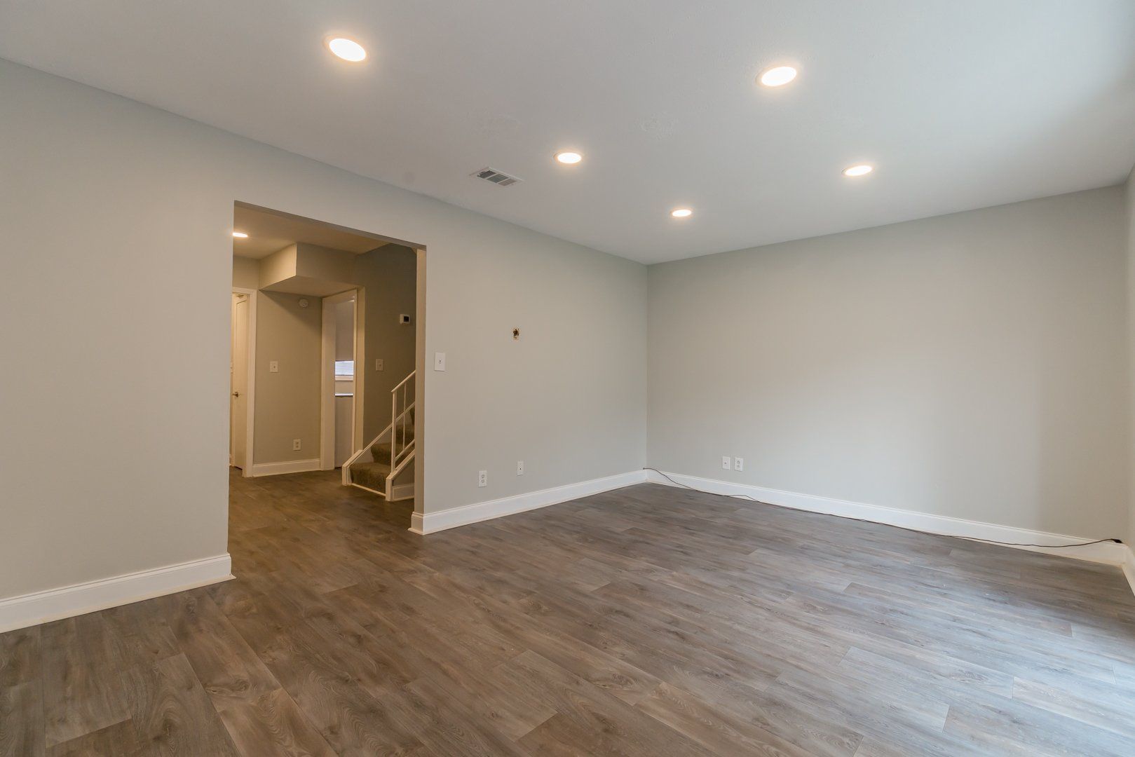Empty modern living room with wood floors, recessed lights, and a doorway to a staircase area