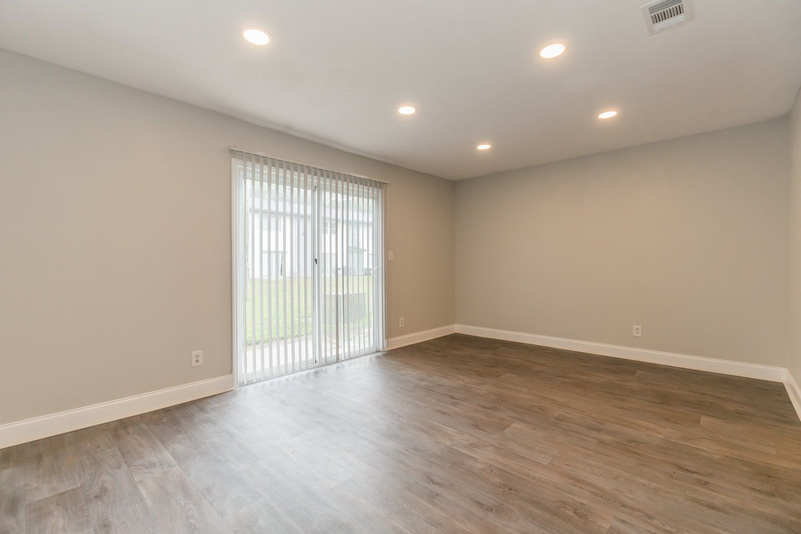 Empty modern living room with gray walls, wood floors, and sliding glass door with vertical blinds