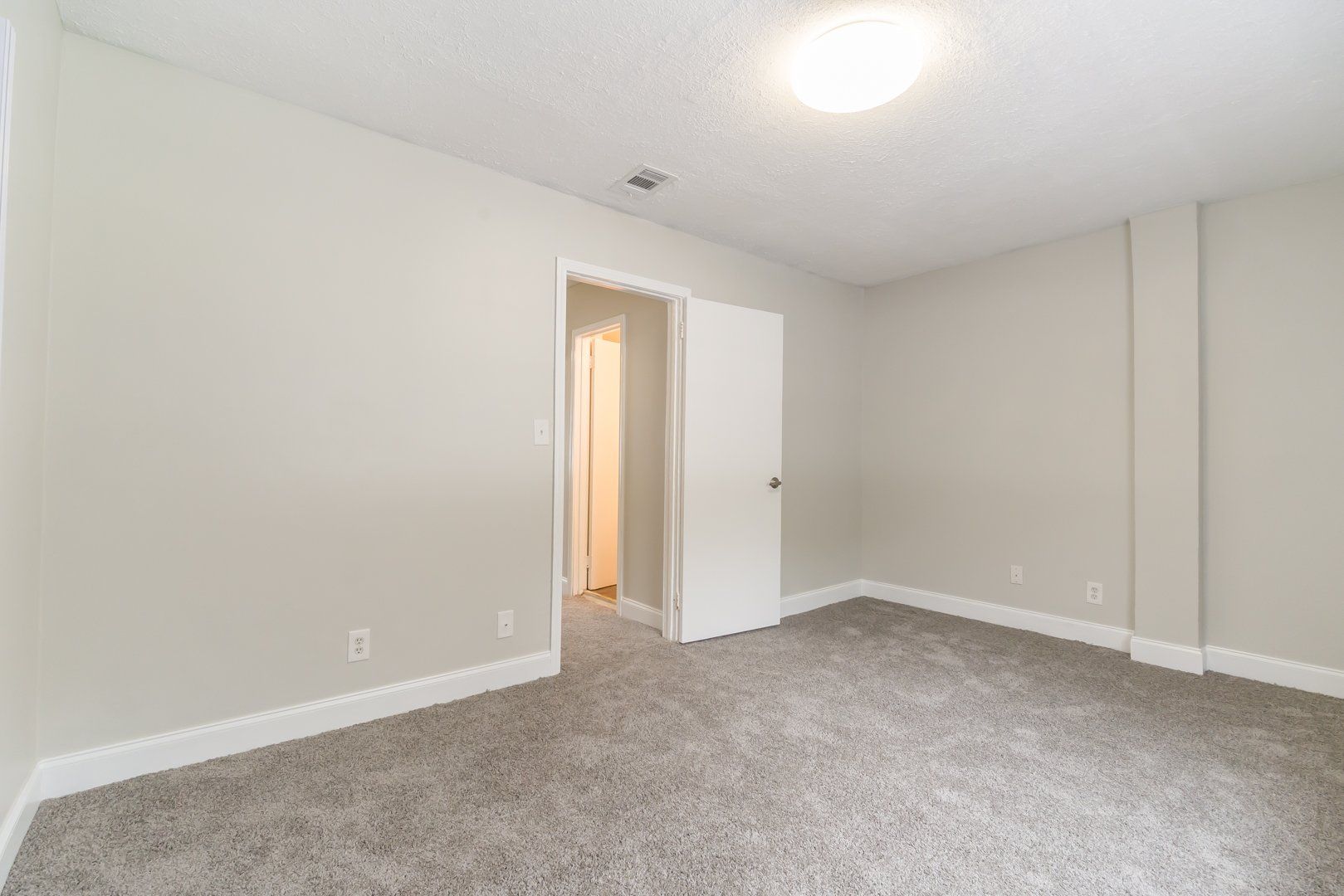 Empty beige bedroom with gray carpet and open door to a lit hallway