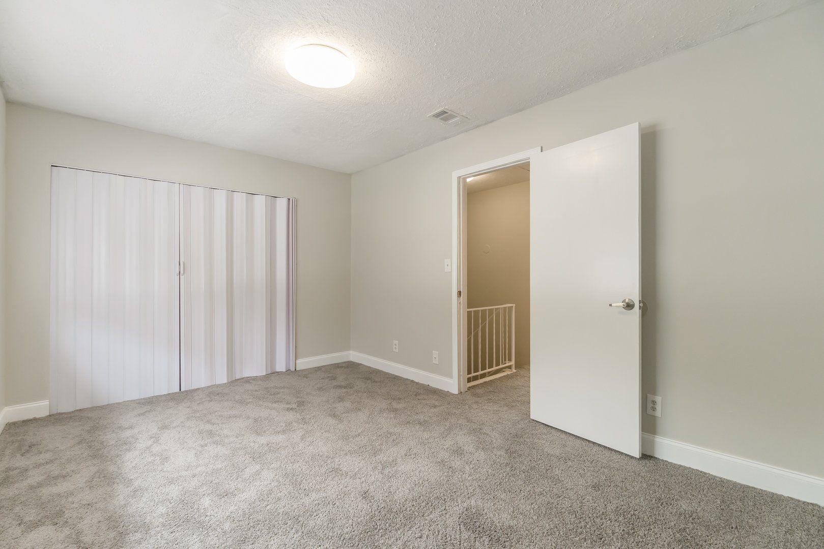 Empty beige bedroom with carpet, closed blinds, and an open door to a hallway