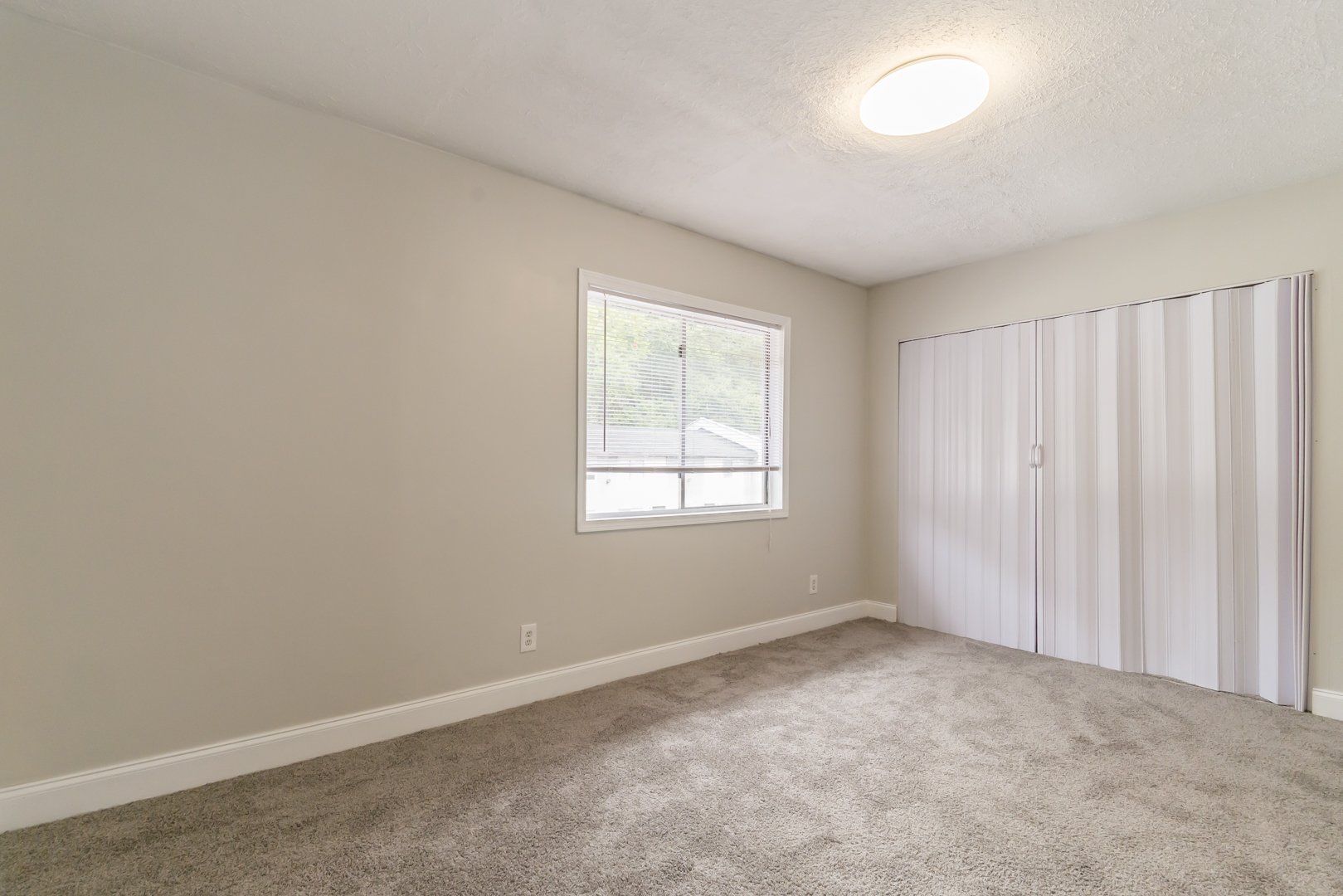 Empty beige bedroom with carpet, ceiling light, window, and sliding closet doors