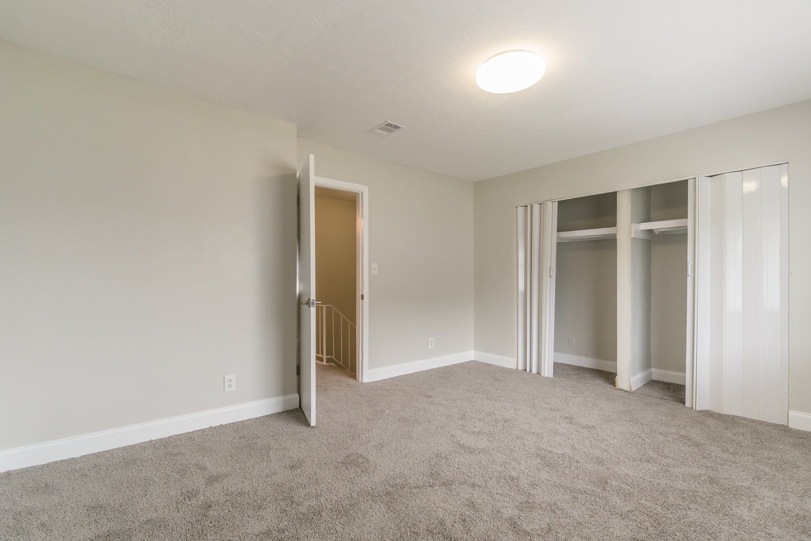 Empty beige bedroom with carpet, open door, and white closet doors
