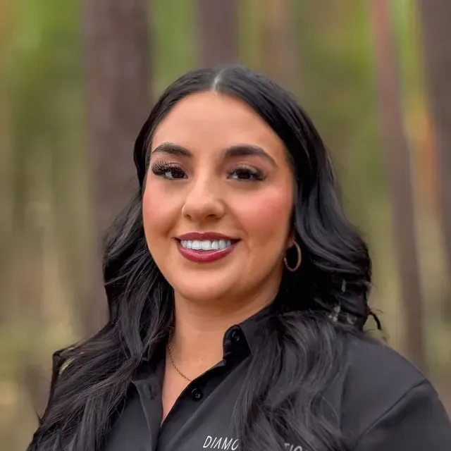 Smiling woman in a black shirt standing outdoors with blurred trees in the background