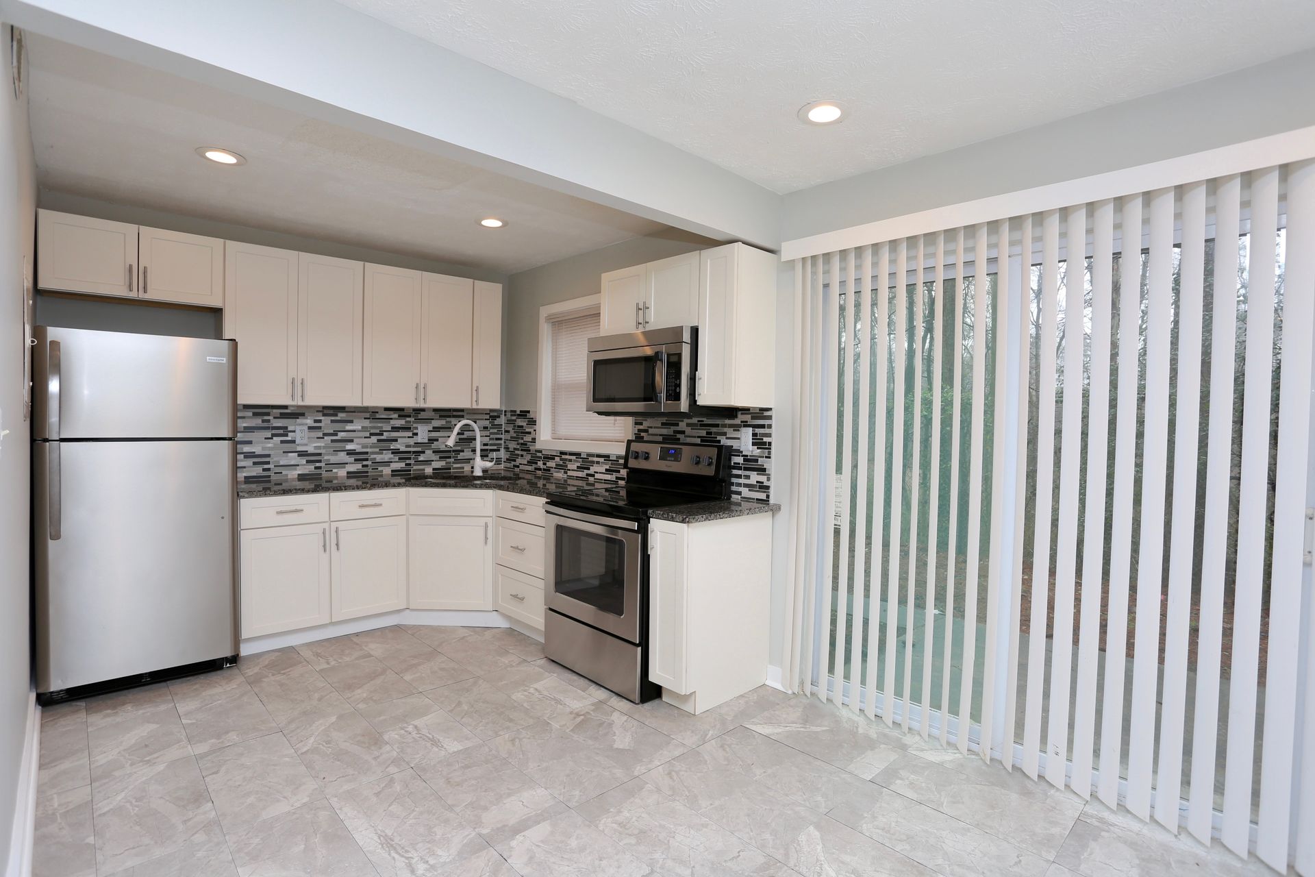 Bright kitchen with white cabinets, stainless fridge, stove, and vertical blinds by a sliding door.