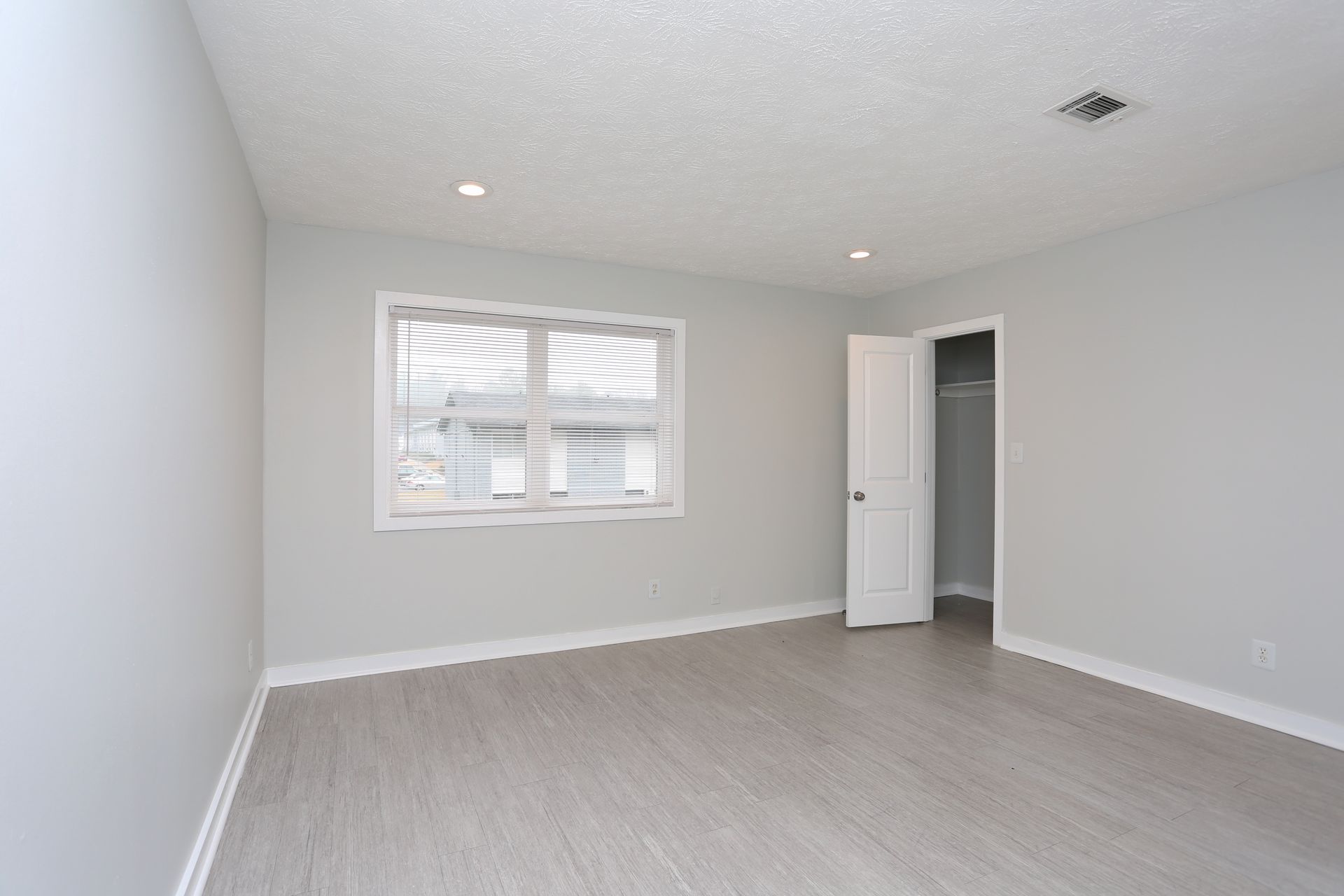 Empty light-gray bedroom with window, white door, and carpeted floor.