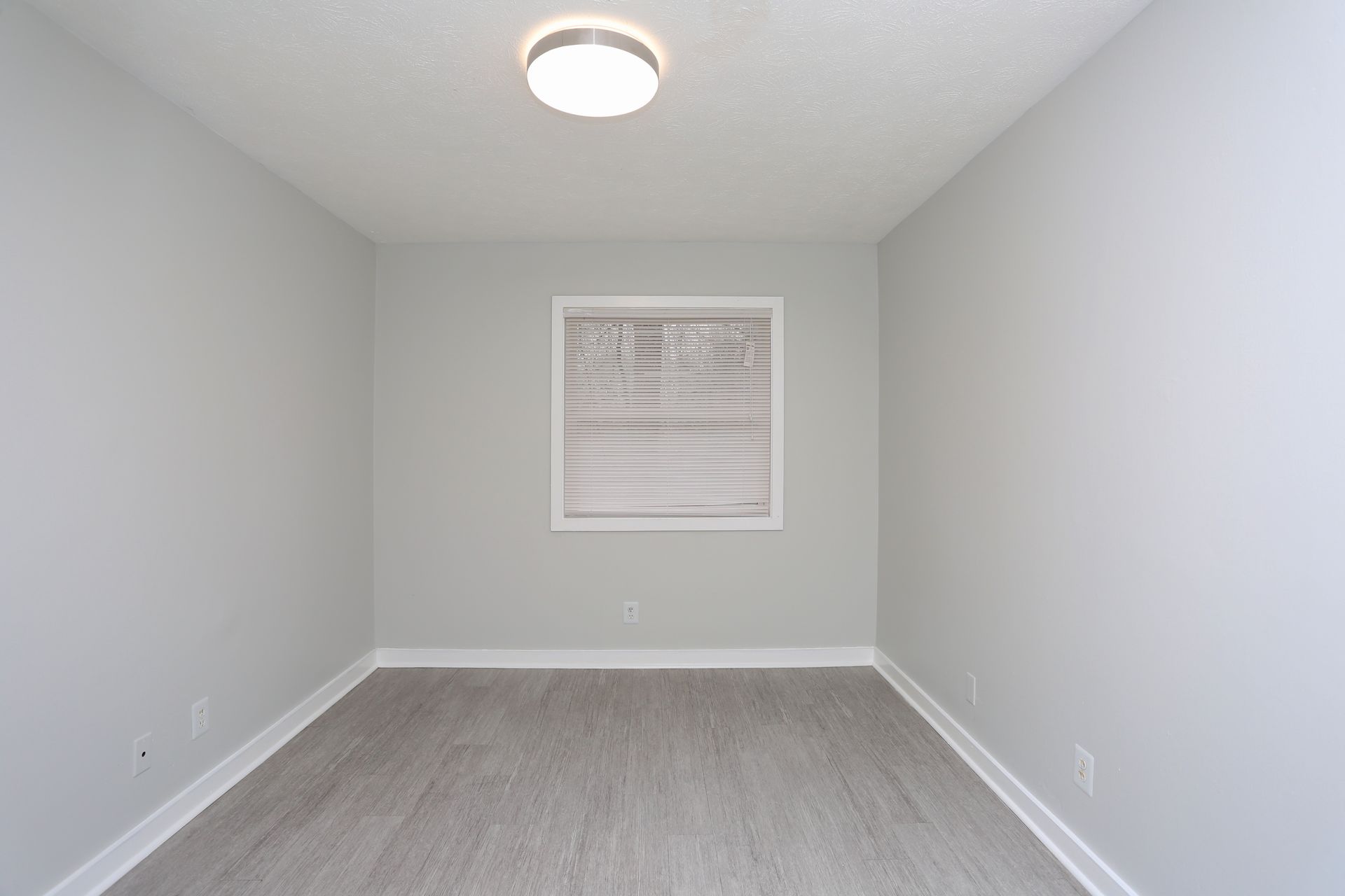 Empty gray room with carpet, a centered window with blinds, and a ceiling light.