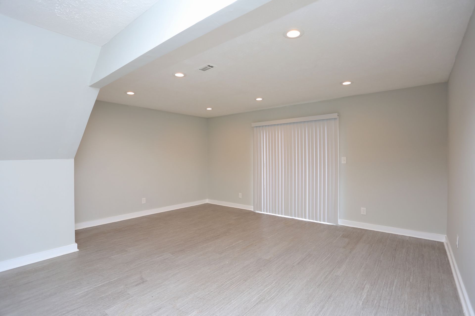Empty beige carpeted room with slanted ceiling, recessed lights, and vertical blinds over a sliding door