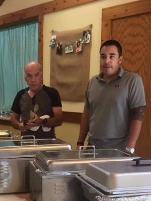 Two men standing behind a buffet table with covered serving trays in a dining hall