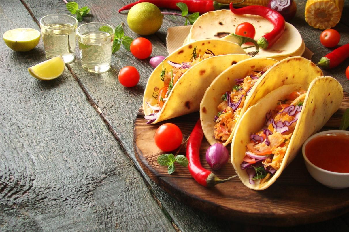 Tacos on a wooden board with limes, tomatoes, and salsa on a rustic table