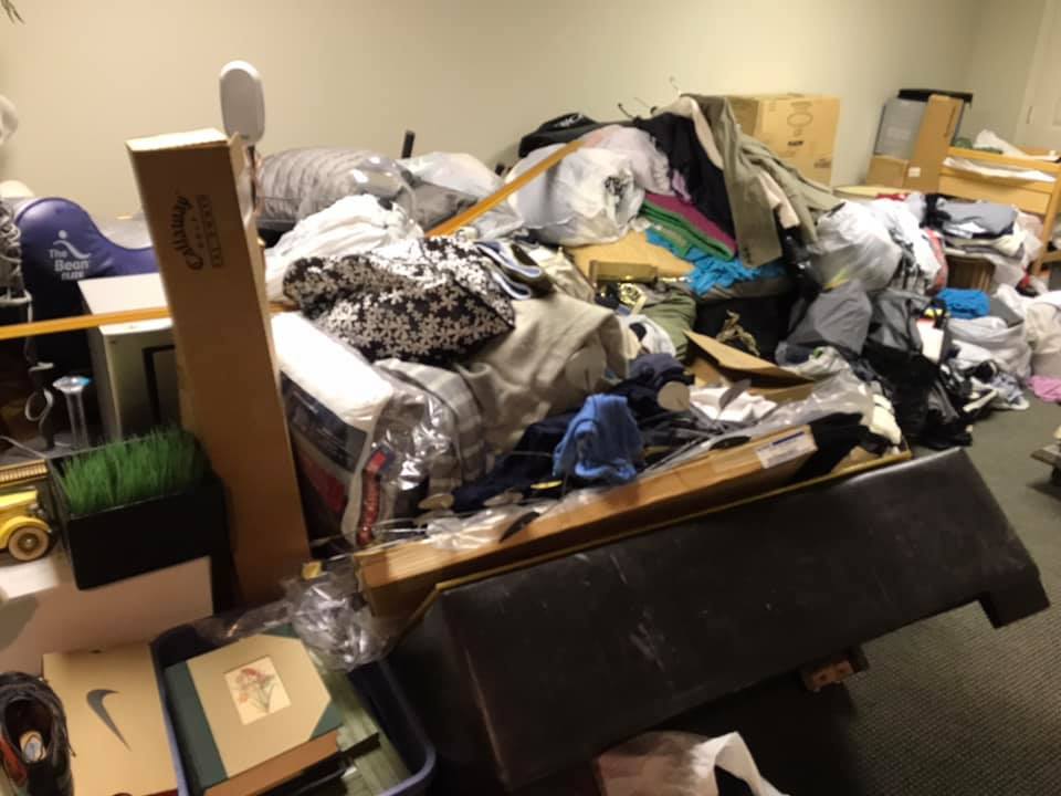 A cluttered room with clothes, boxes, and furniture piled high.