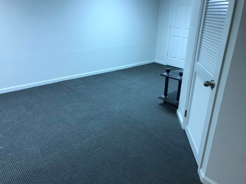 Empty room with blue-gray carpet, white walls, and a closed door. A small black table sits in the corner.