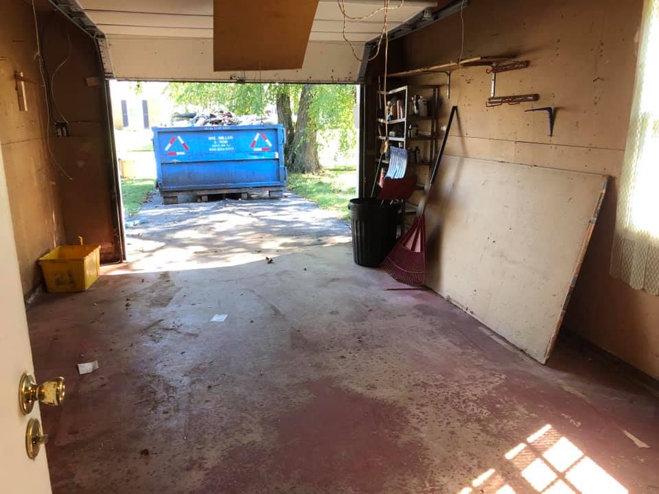 Inside a messy garage, a dumpster is visible through the open door.