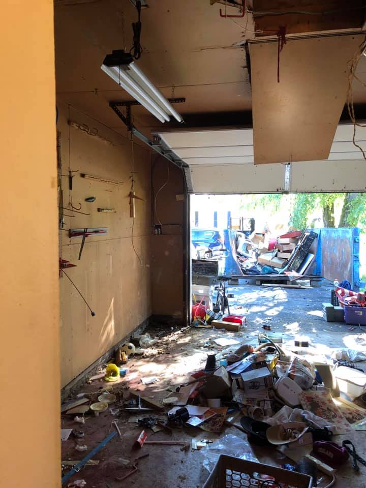 Interior view of a messy, destroyed garage with debris on the floor and an open doorway to an outdoor area.