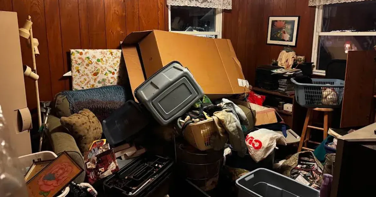 Cluttered room with piled cardboard boxes, bins, and assorted household items against wood-paneled walls.