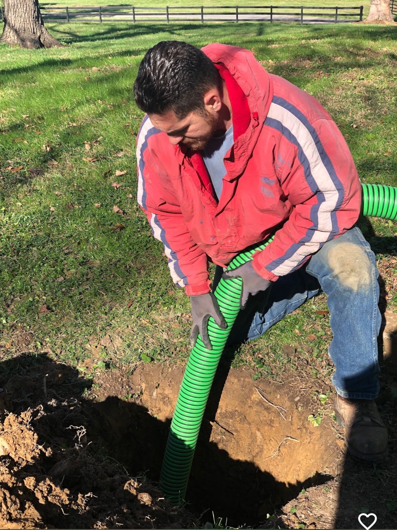 Man in red jacket digging a hole with a green shovel in a grassy yard