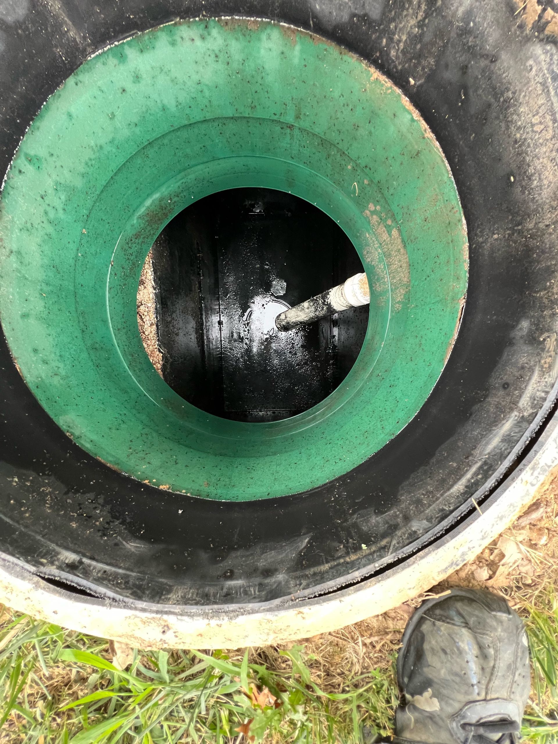 Close-up of a green-rimmed circular pipe opening in the ground beside a boot and grass.