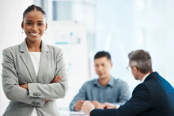 Professional standing in a modern office with colleagues collaborating in the background.