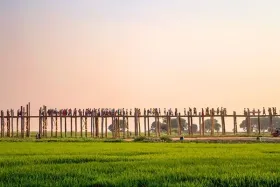 People walking across a long wooden bridge above green fields under a clear evening sky.