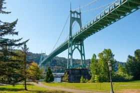 Suspension bridge spanning a river, viewed from a green park with trees under a clear blue sky.