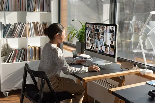 Person participating in a video conference on a desktop computer in a bright home office or workspace.