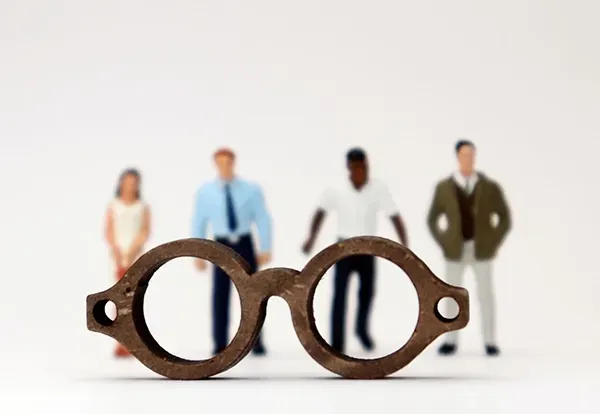Pair of eyeglasses in the foreground with blurred miniature figures in the background, symbolizing perspective and awareness.