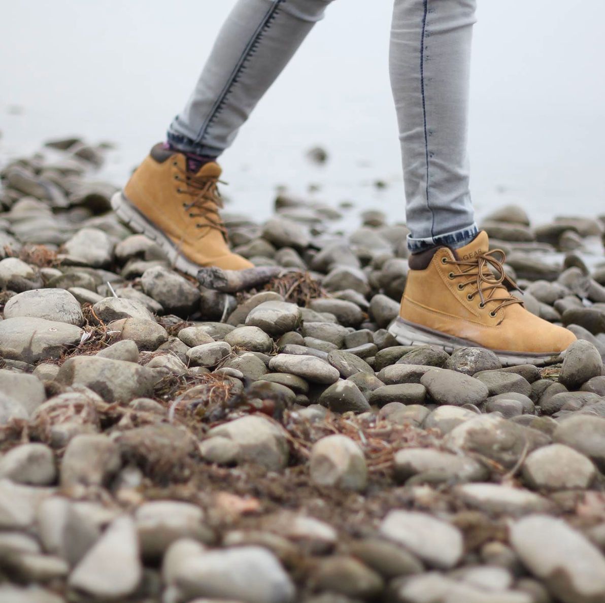 Tan boots and gray jeans stepping across a rocky beach shoreline