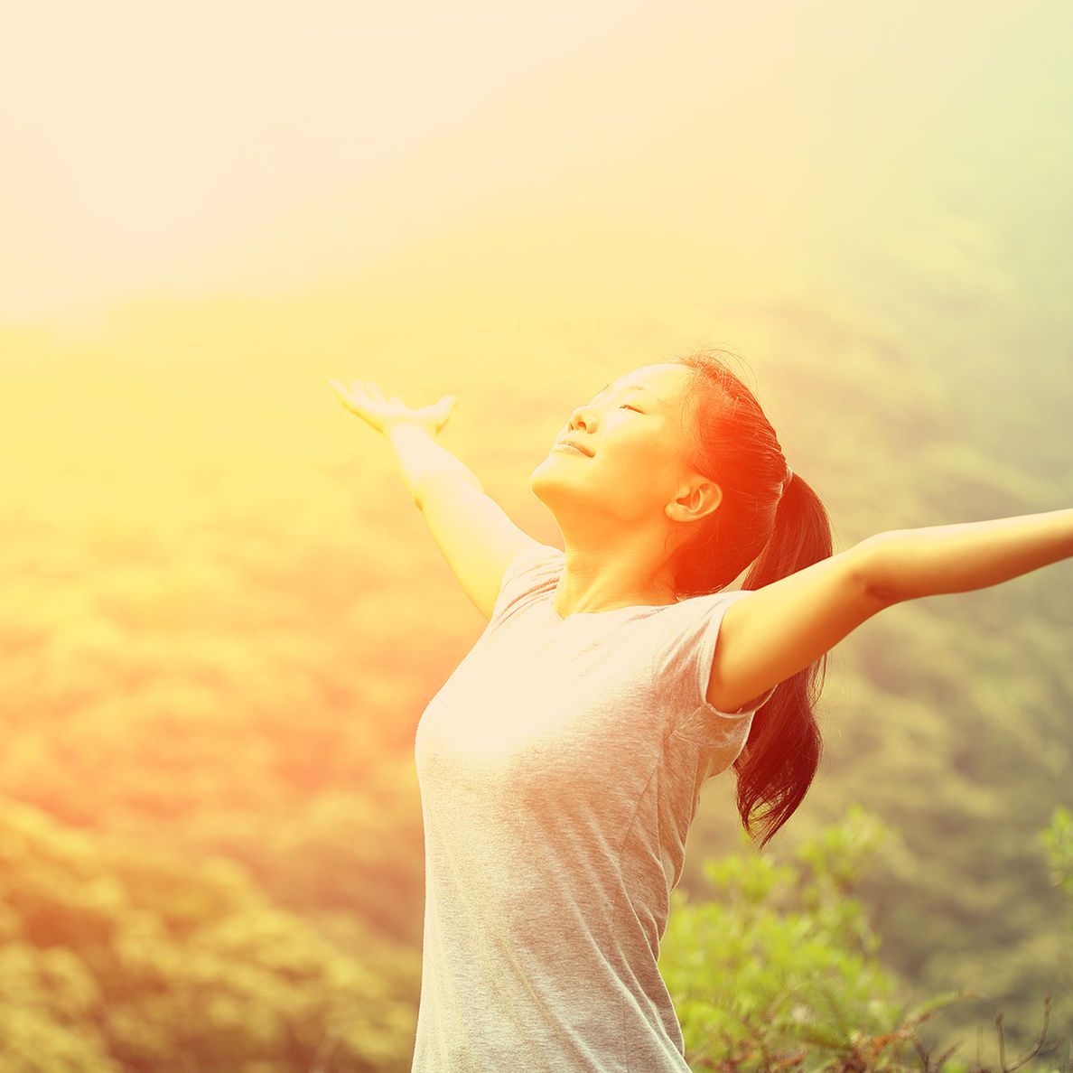 Woman standing outdoors with arms outstretched in warm sunlight over a green landscape