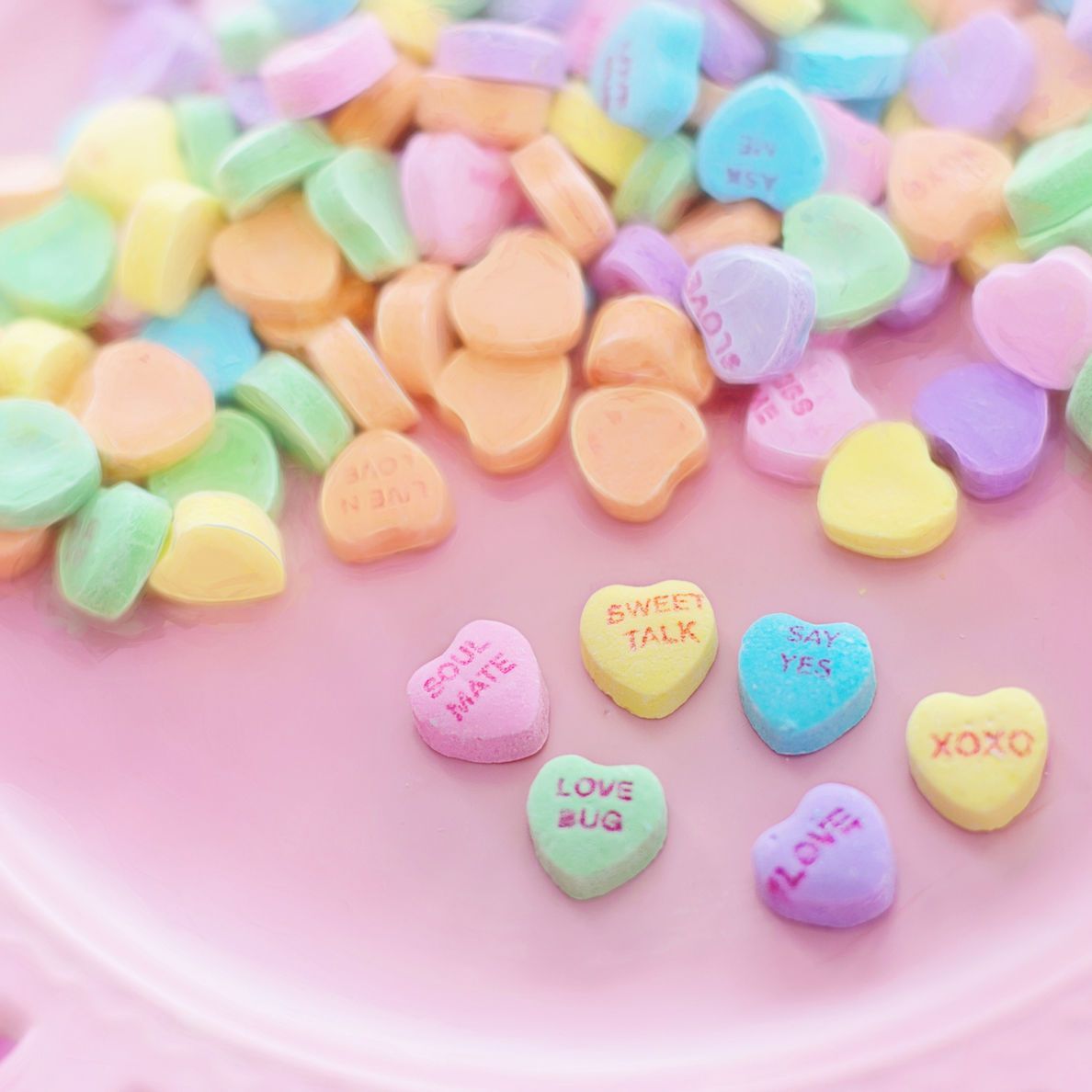 Pastel conversation heart candies scattered in a pink bowl, with a few hearts reading “Love You” and “XOXO.”