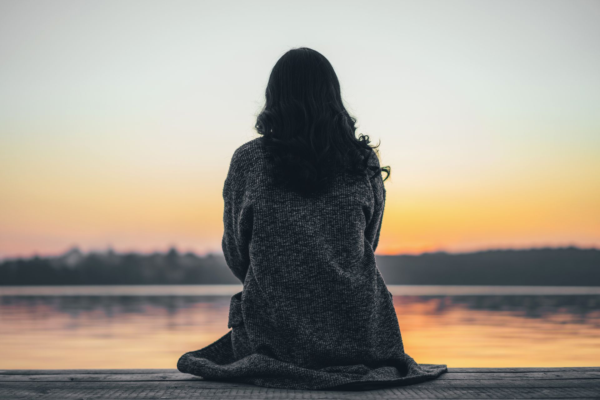 Person seated on a dock at sunset, facing a calm lake with an orange horizon