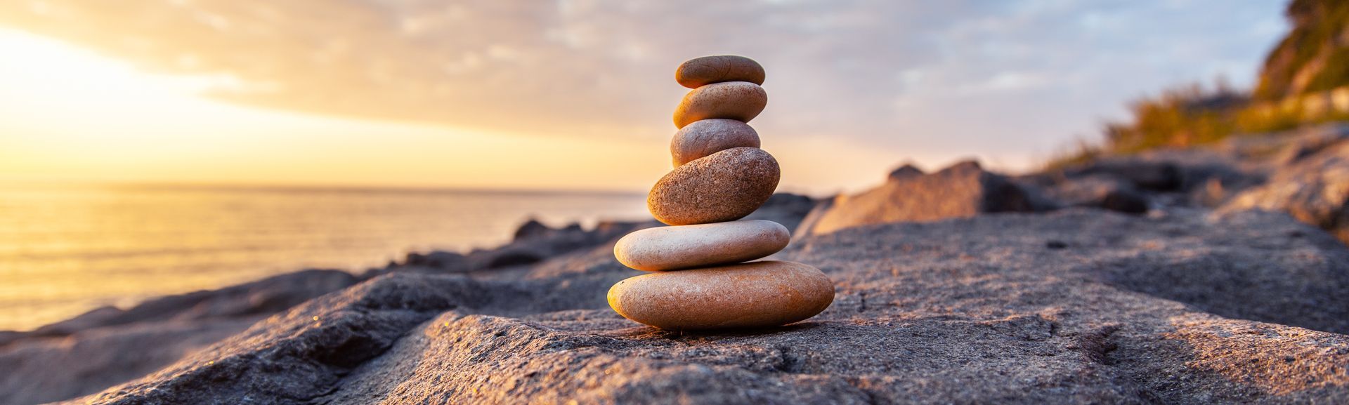 Stacked smooth stones on a sandy beach at sunset, with warm orange light and a calm sea in the background