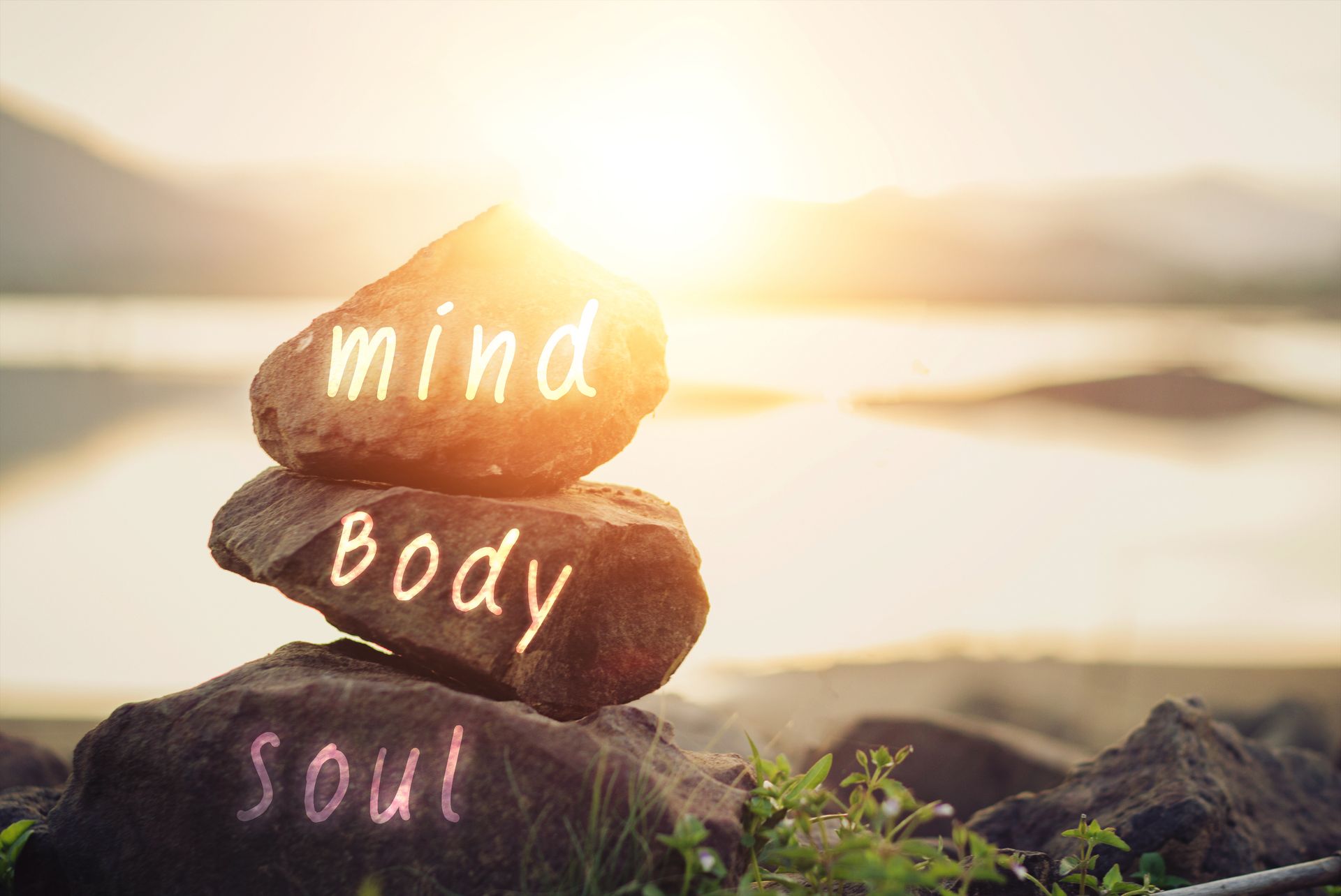 Stacked stones labeled mind, body, soul by a sunlit lake at sunrise