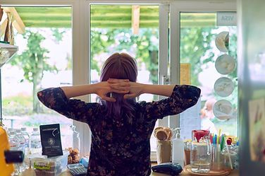 Person with arms raised stands at a sunlit vanity by large windows, surrounded by makeup and jars.