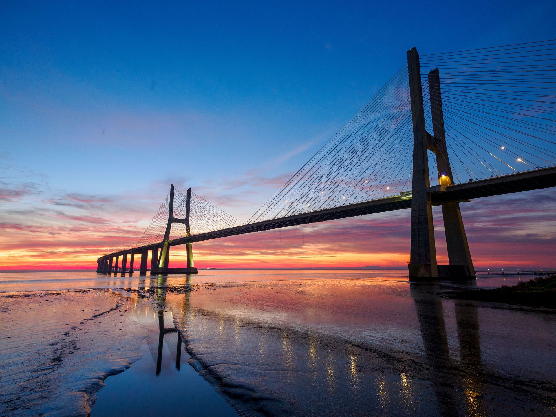Sunset-lit cable-stayed bridge reflecting on calm water under a pink and blue sky