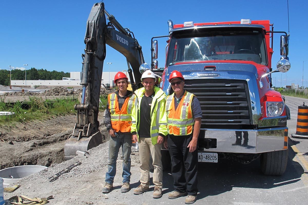 Three construction workers standing in front of a red truck and excavator on a sunny day.
