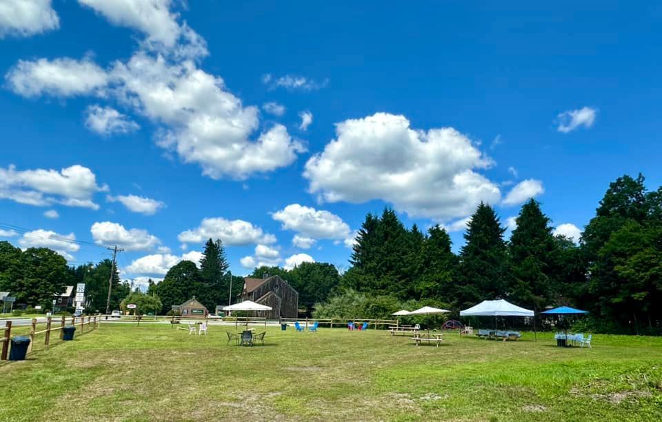Open grassy field with picnic tables, tents, and trees under a blue sky with puffy white clouds.