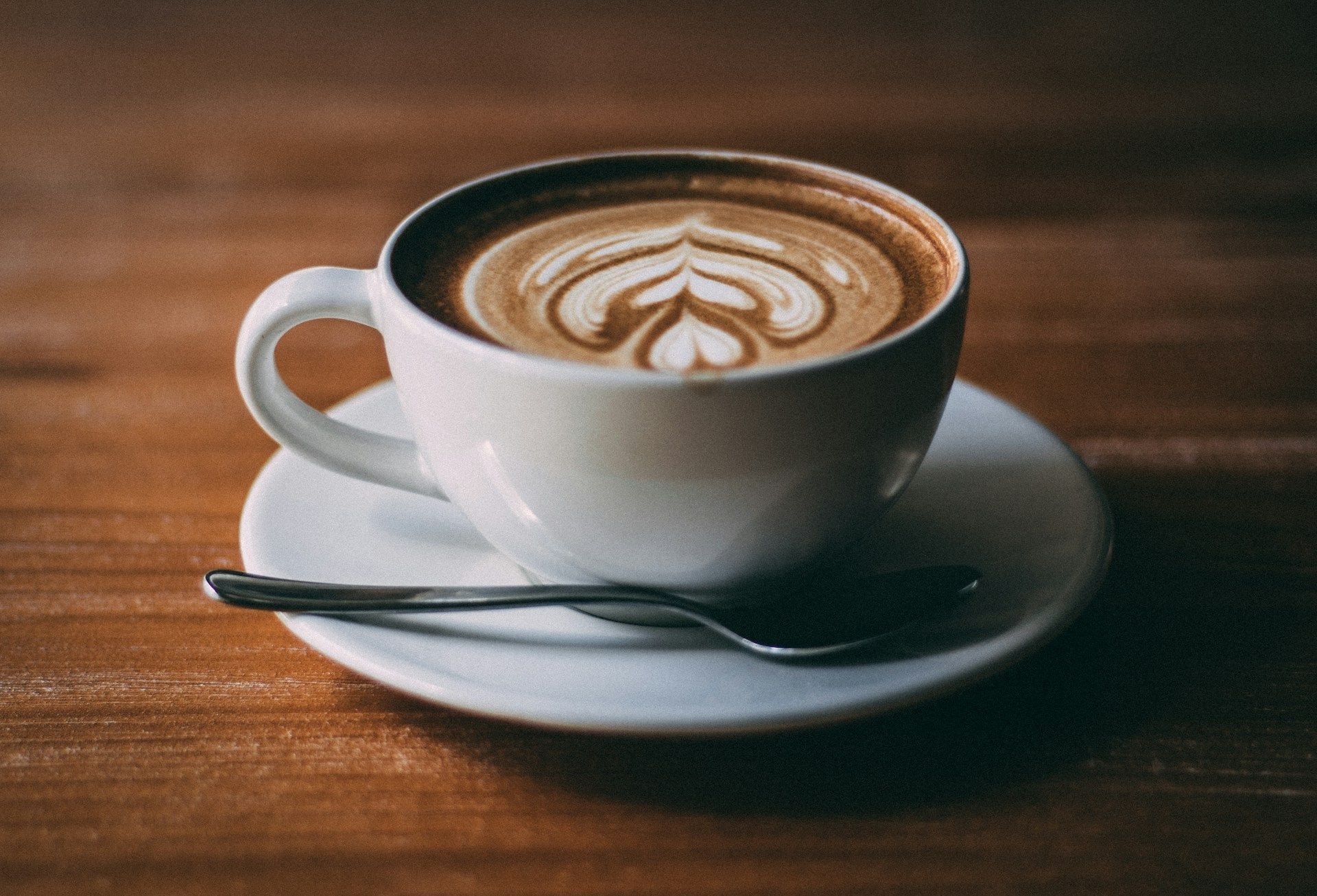 White cup of latte with heart-shaped foam art on a saucer and spoon, on a wooden table