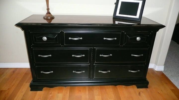 Black dresser with multiple drawers on hardwood floor, with a lamp and small framed screen on top.