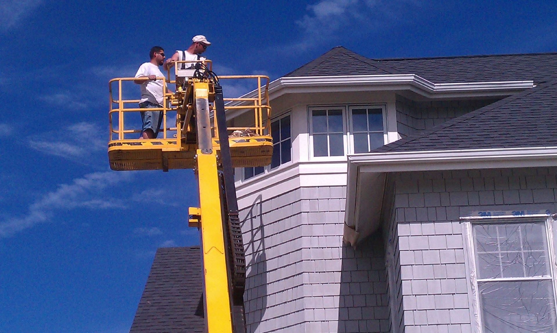 Workers in a yellow lift painting a gray house roofline against a blue sky