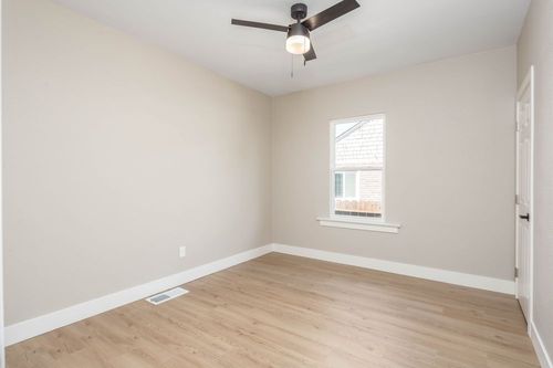 Empty beige bedroom with light wood floors, ceiling fan, and a small window with blinds.