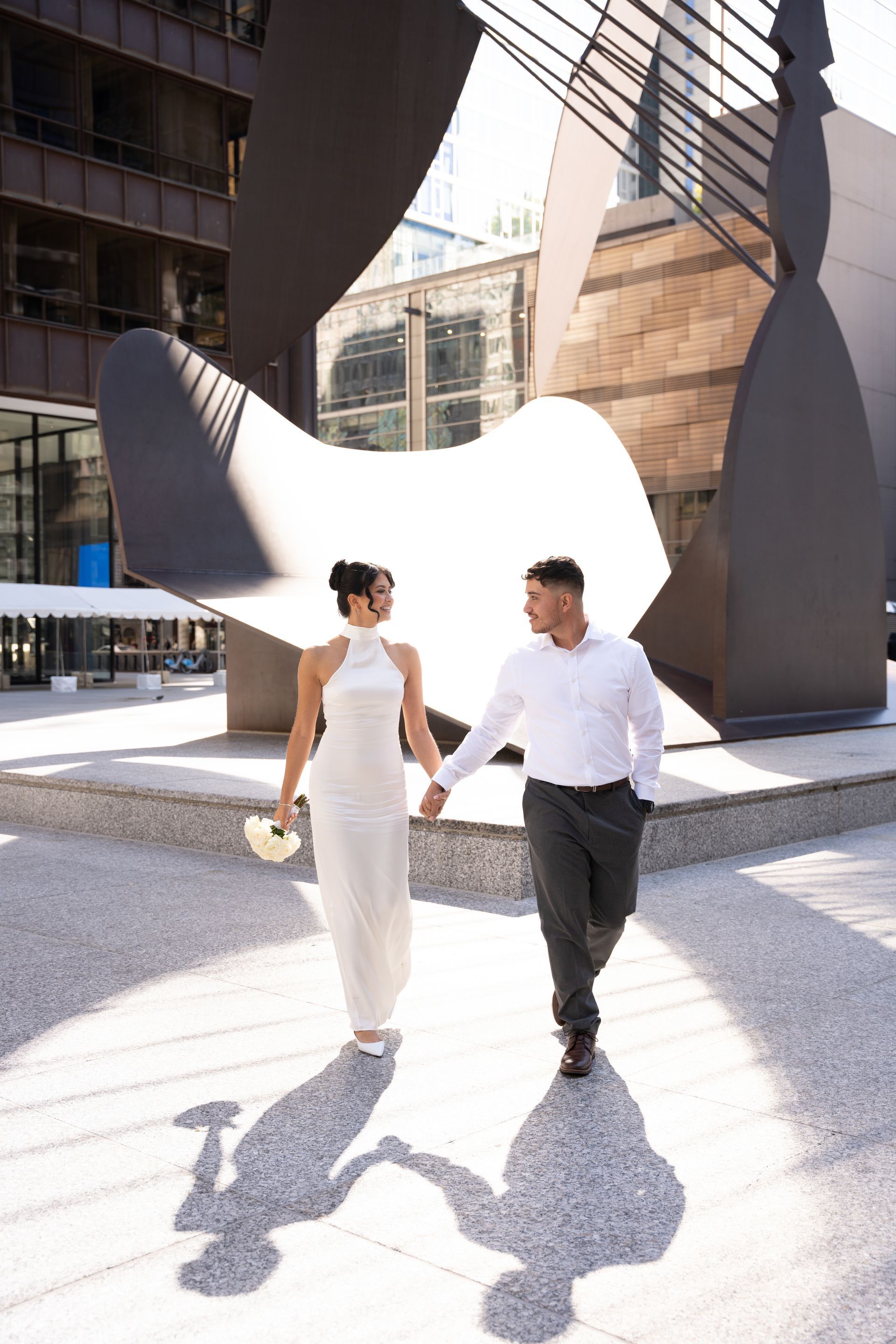 Couple in wedding attire hold hands, walking towards camera near modern sculpture.