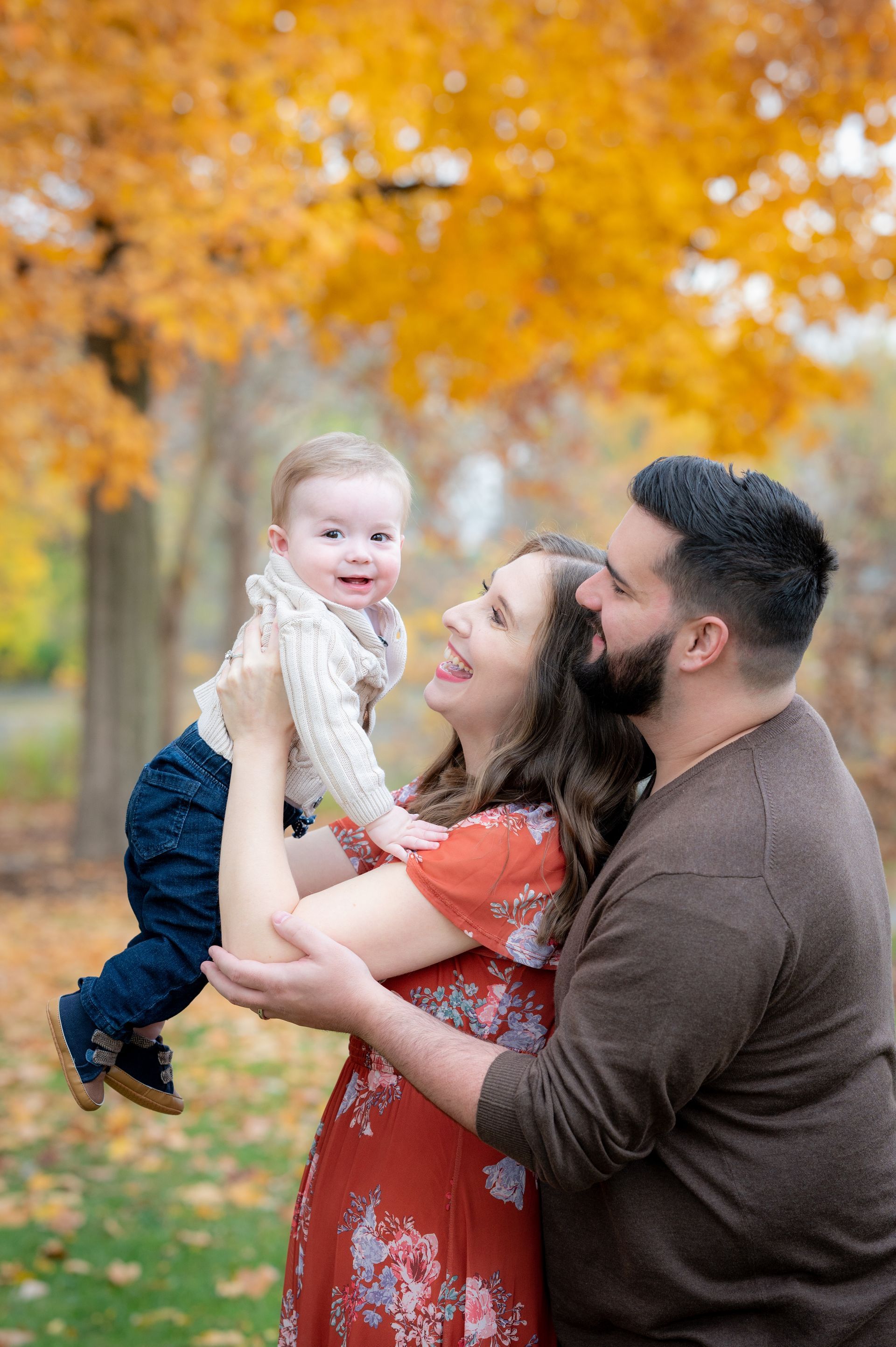 Family of three smiles while playing, under autumn leaves.