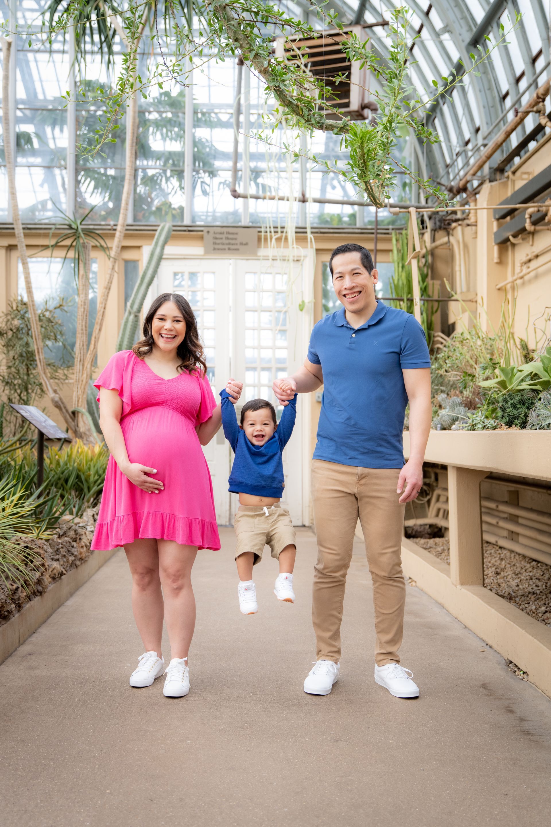 Family in a greenhouse: pregnant woman in pink, child in the air, man in blue. Lush plants, glass walls.