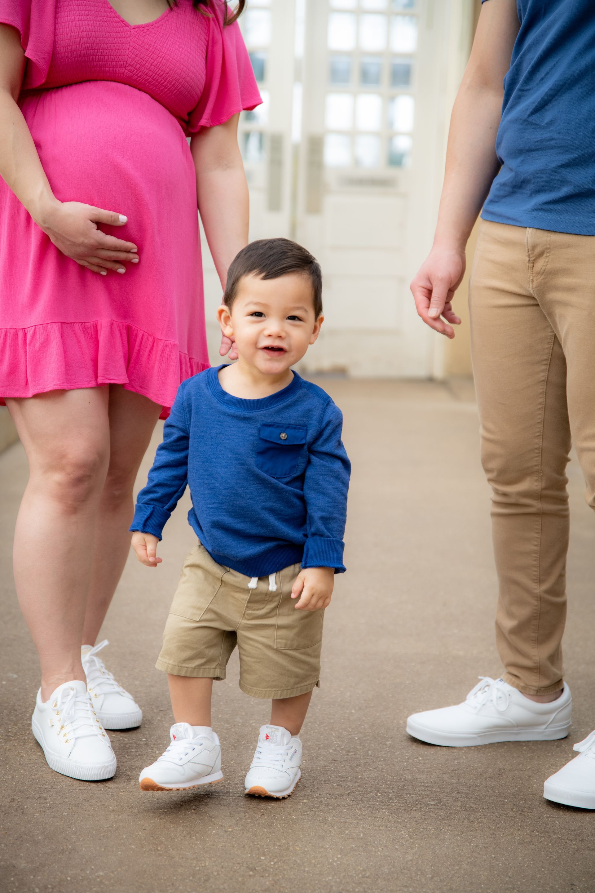 A toddler smiles, standing between two adults; one pregnant, all wearing casual clothes. Outdoors.
