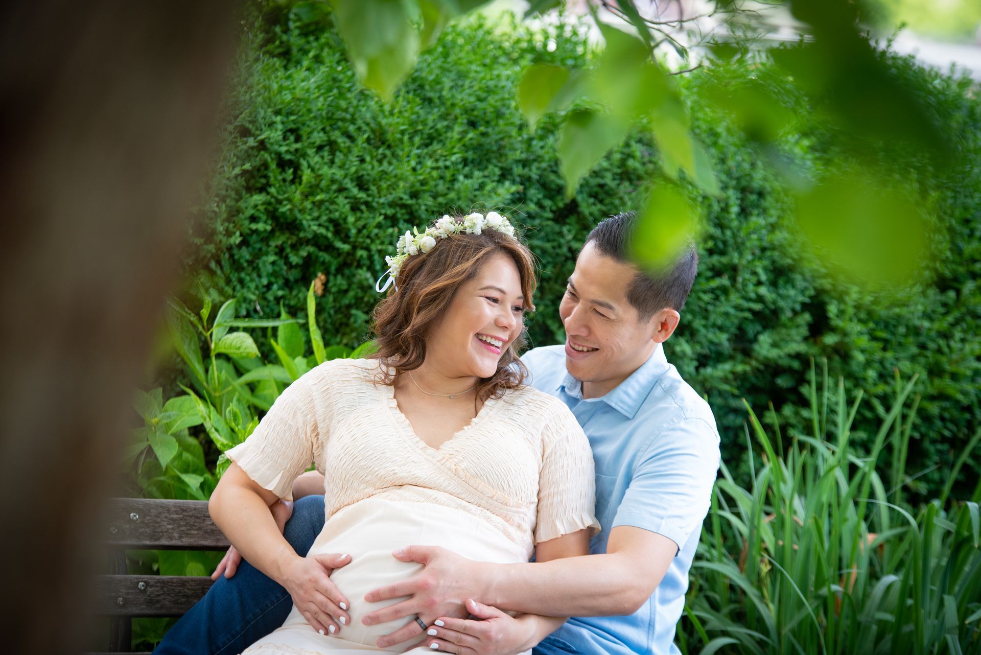Pregnant couple sits on bench, smiling and holding belly in a garden.